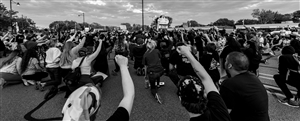 A black and white photo of a crowd of people holding their fists above their heads, looking at a point on the horizon. Photo by  Cooper Baumgartner via Unsplash.